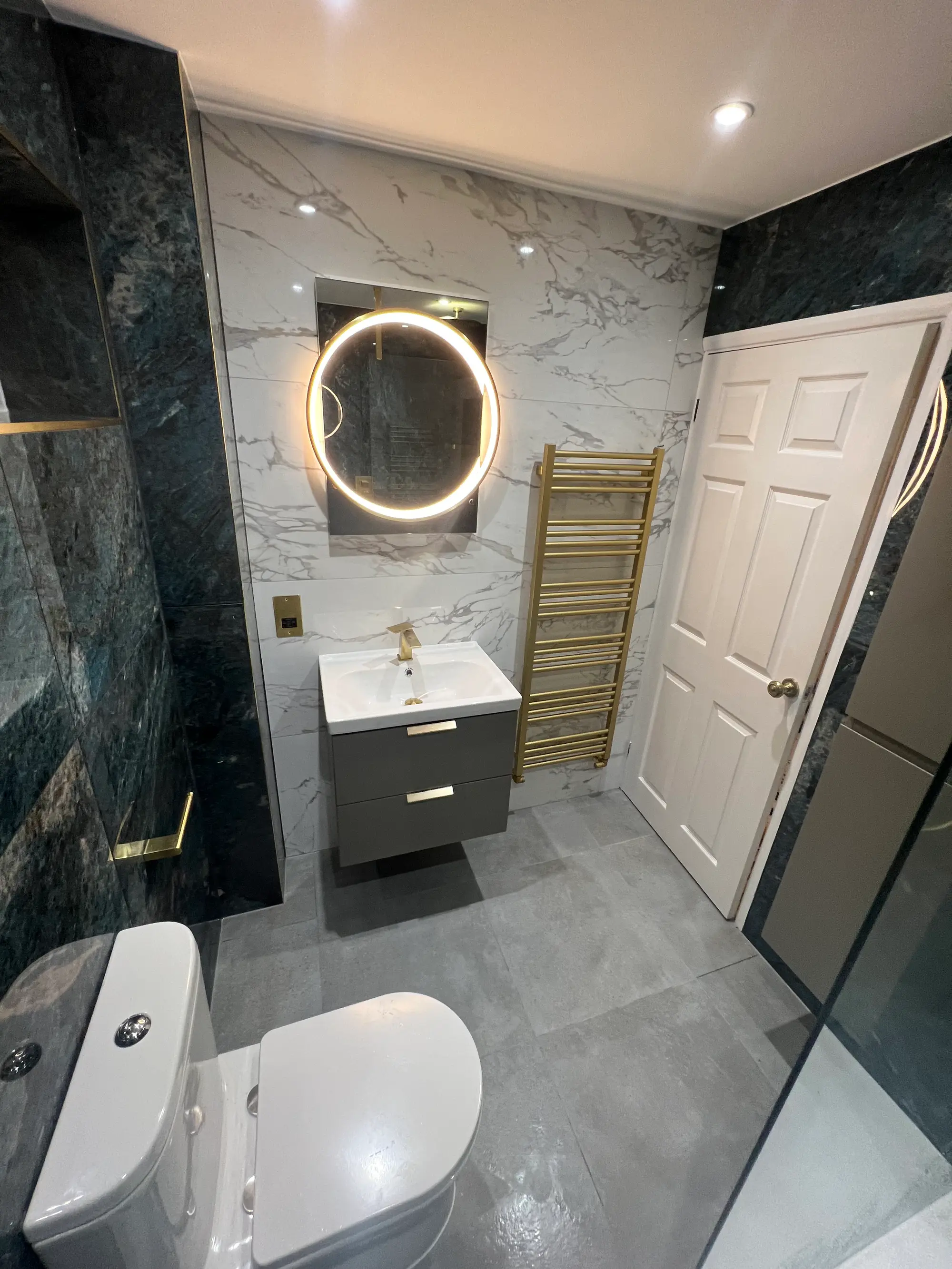 Green marble bathroom with brass fittings and an illuminated round mirror